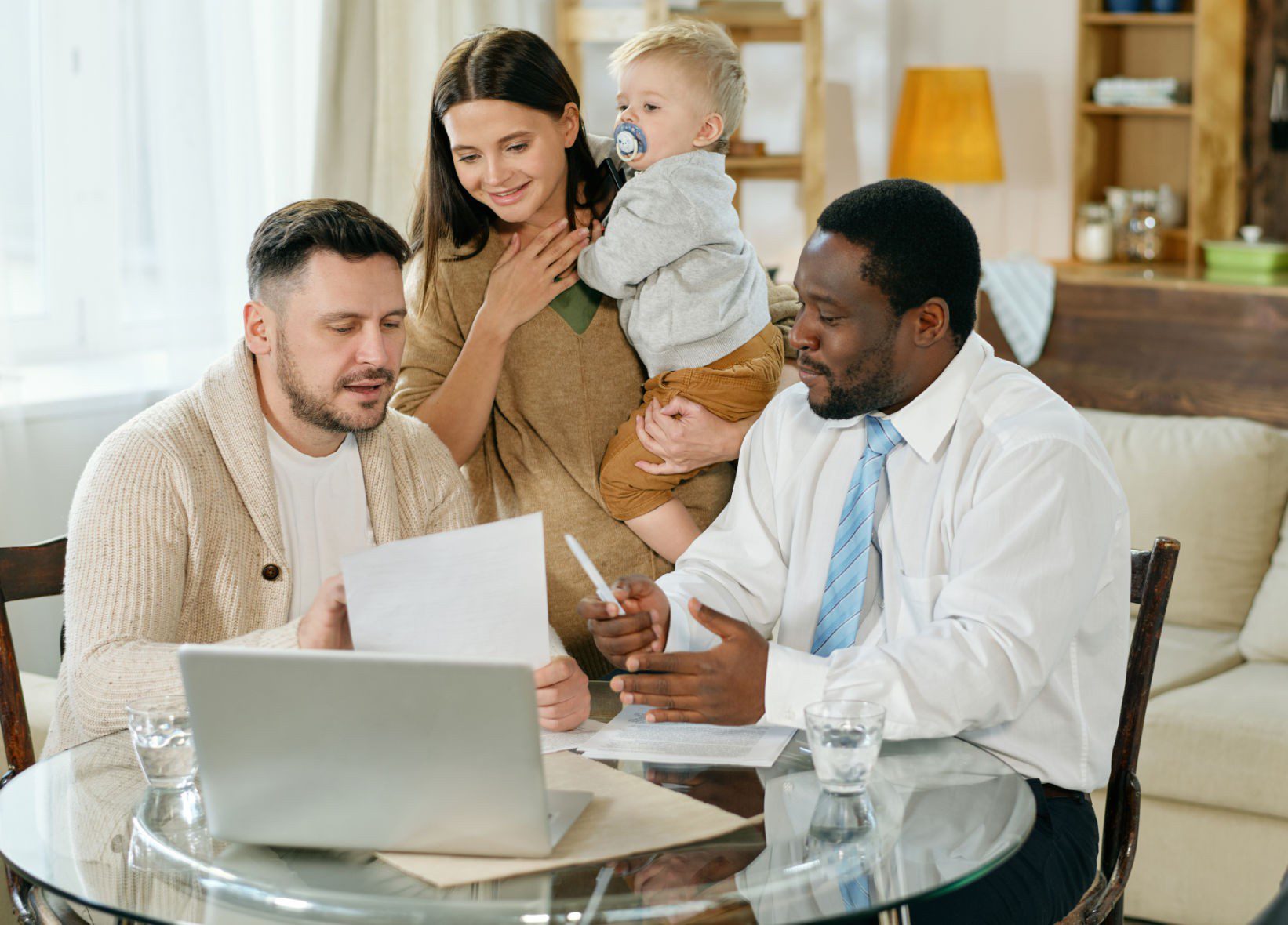 A couple with a young child consults with a professional at a table, reviewing documents and using a laptop in a well-lit living room.