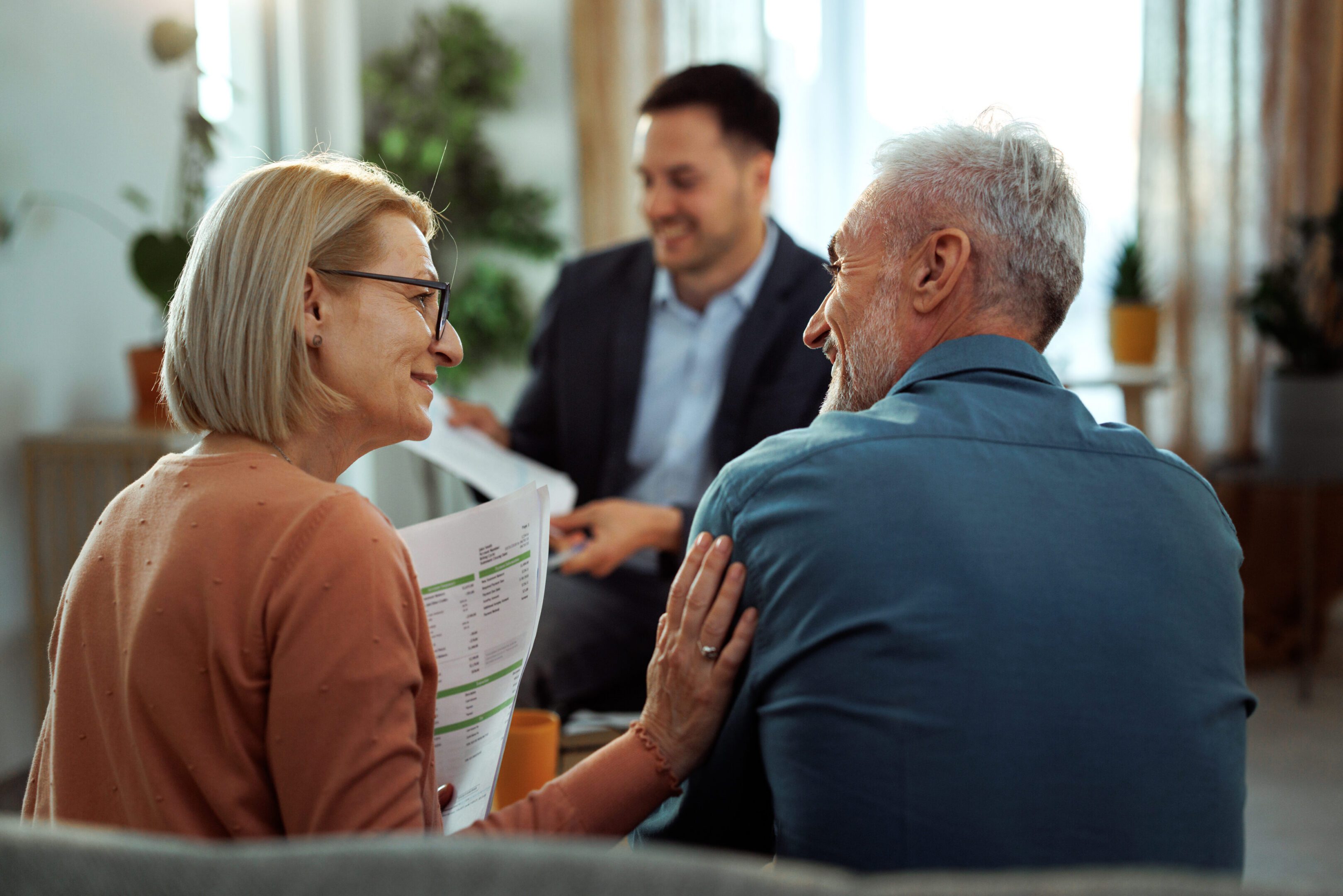 An older couple sits together, holding paperwork and smiling, while a man in a suit sits across from them in a bright room with plants.