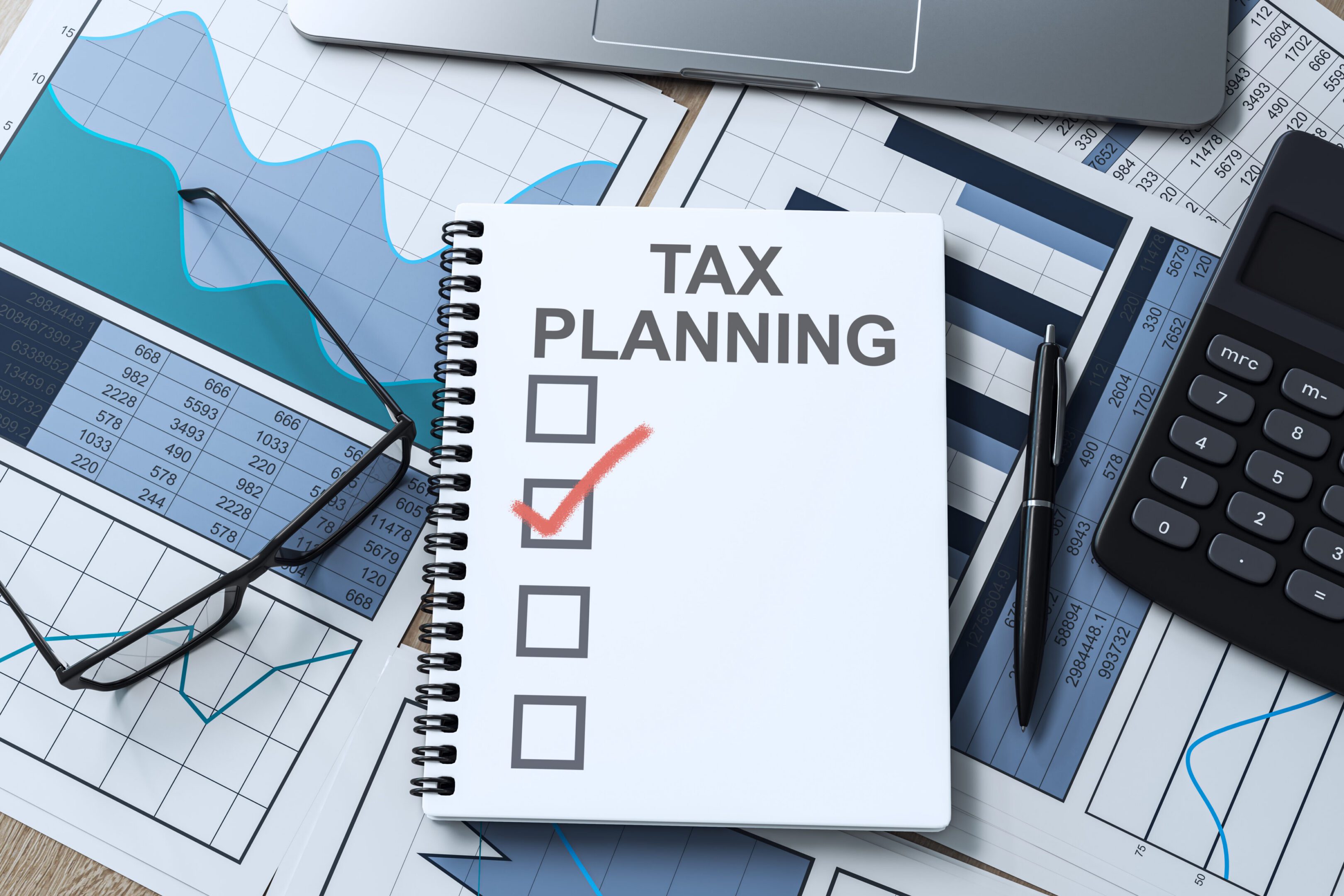 A notebook labeled "Tax Planning" with a checked box, surrounded by a calculator, glasses, pen, laptop, and financial charts.