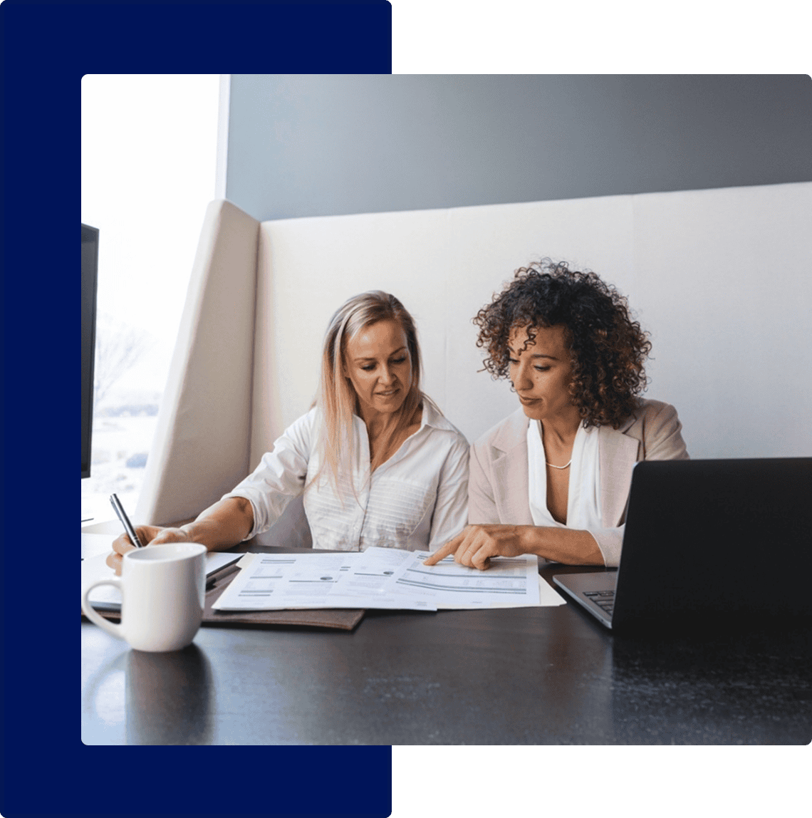 Two women sit at a table with documents and a laptop, discussing paperwork. One is taking notes while the other points at a paper. A coffee mug sits nearby.