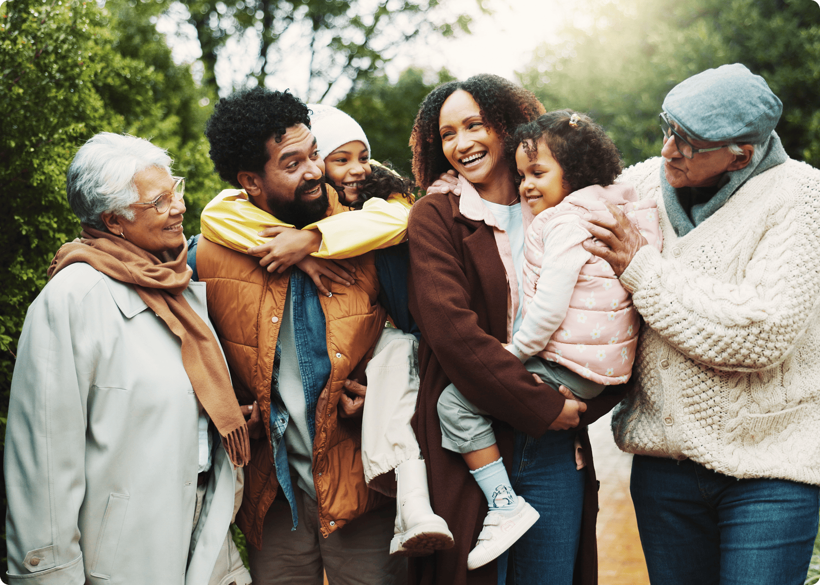 Three adults, two children, and one elderly woman stand outdoors, smiling and interacting with each other, surrounded by greenery.