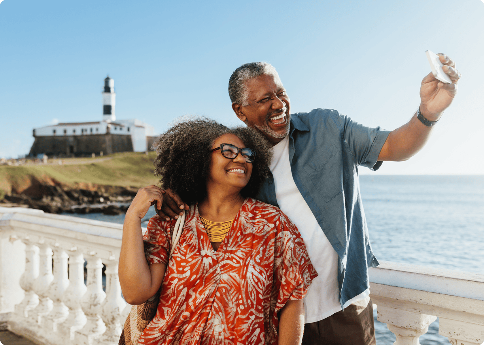 Older couple smiling and taking a selfie near a waterfront, with a lighthouse and grassy hill visible in the background on a sunny day.