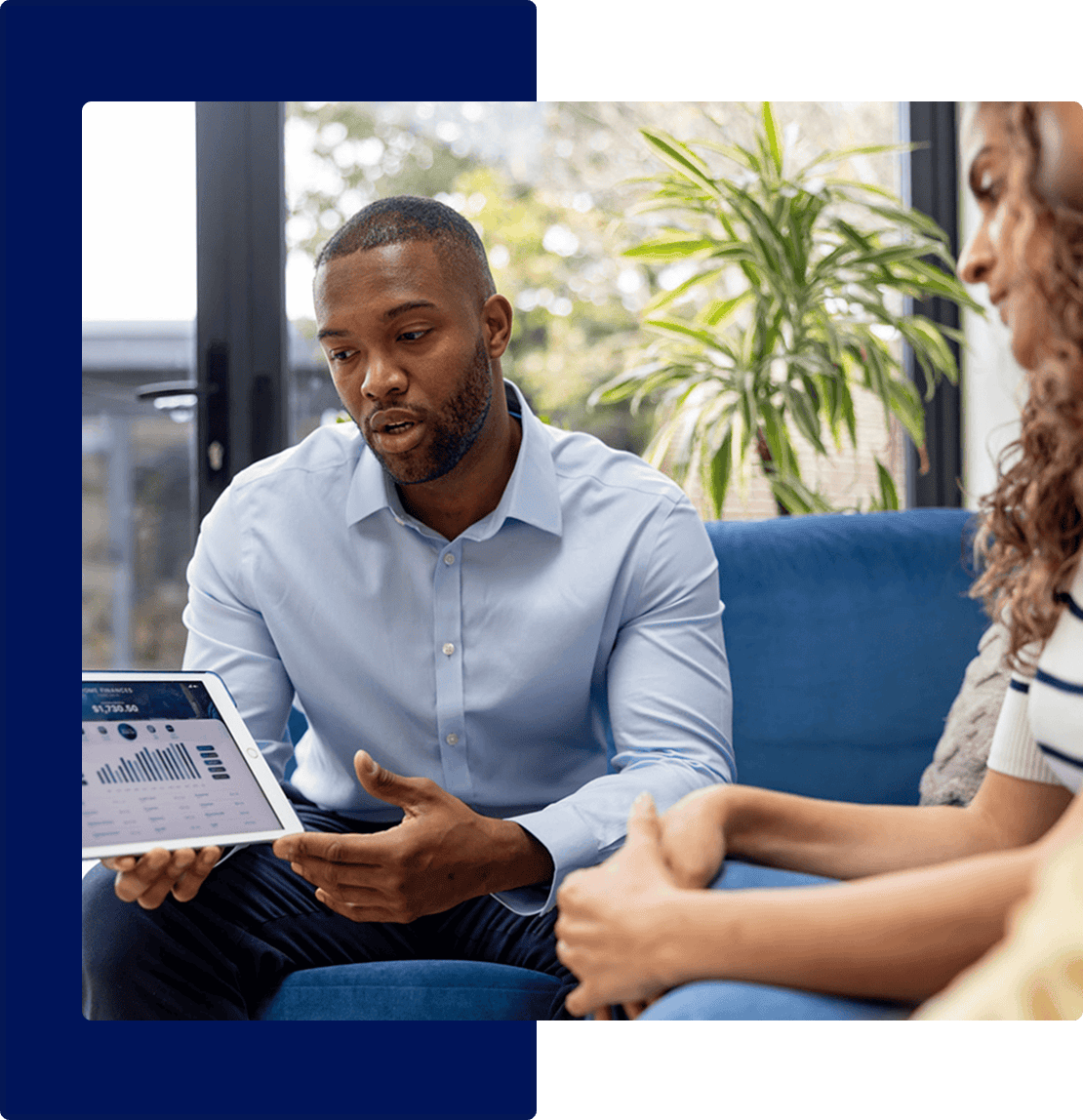 A man in a light blue shirt shows a tablet with a bar chart to a woman on a blue couch in a modern, well-lit room with a plant in the background.