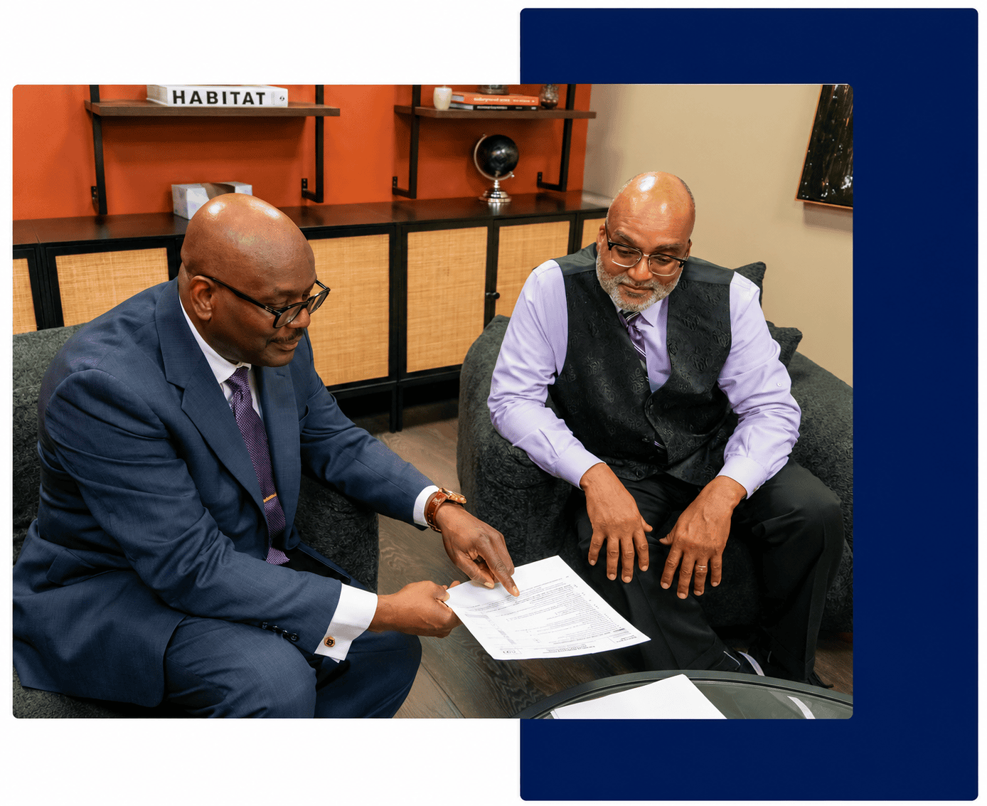 Two men in business attire sit in an office, reviewing a document together. One man points to the paper while the other listens attentively.