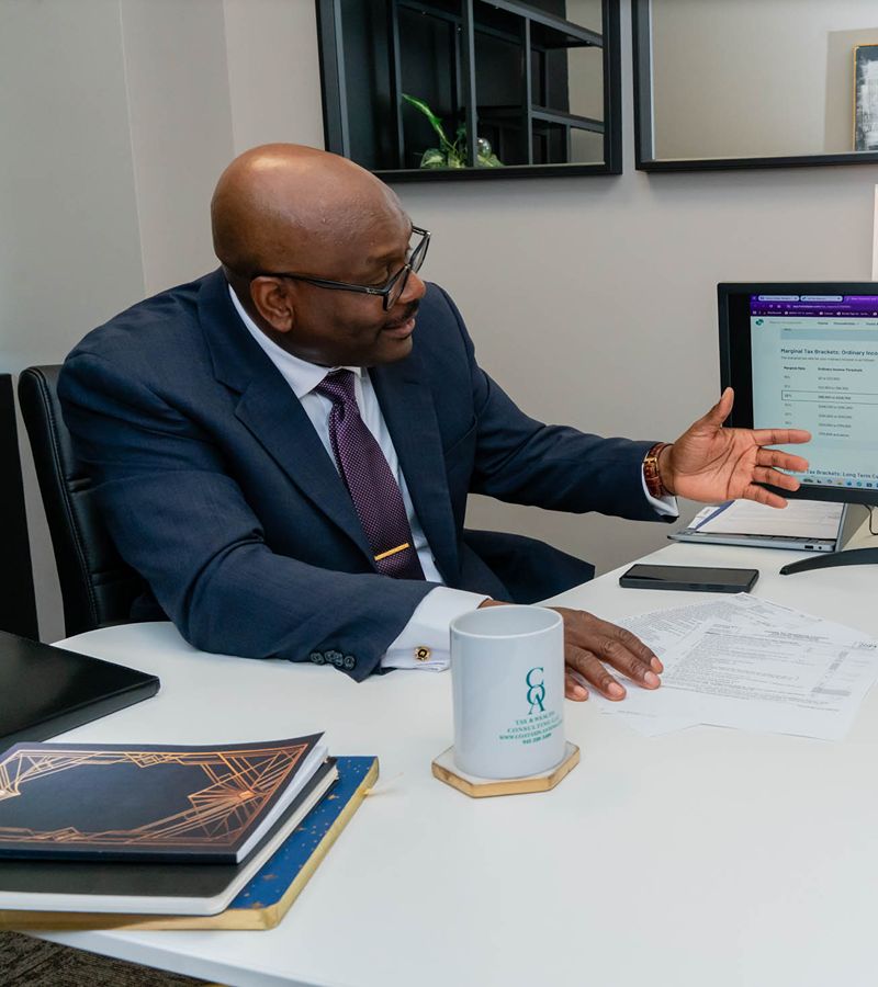 A man in a suit sits at a desk with documents, a computer, a notebook, and a mug, gesturing with his right hand during a conversation.