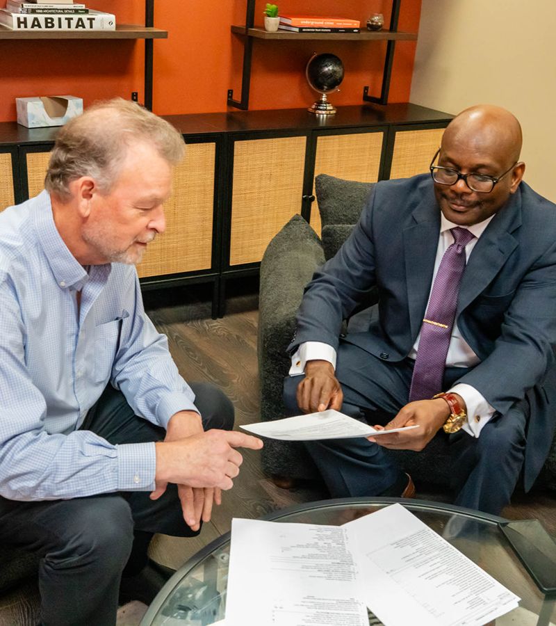 Two men sit in an office discussing documents on a glass table, with shelves and office decor visible in the background.