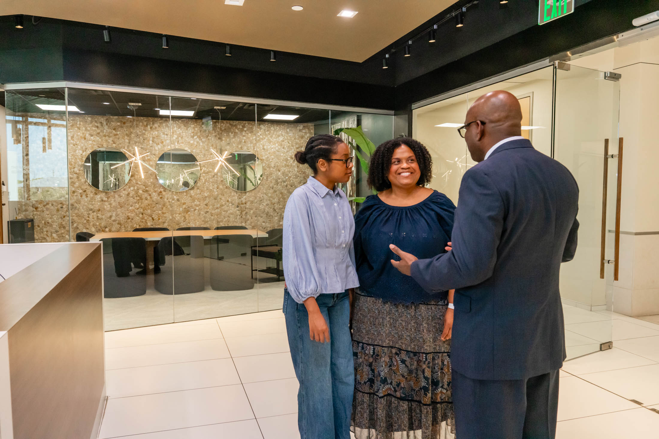 A man in a suit talks to two women standing in a modern office lobby with glass walls and contemporary decor.
