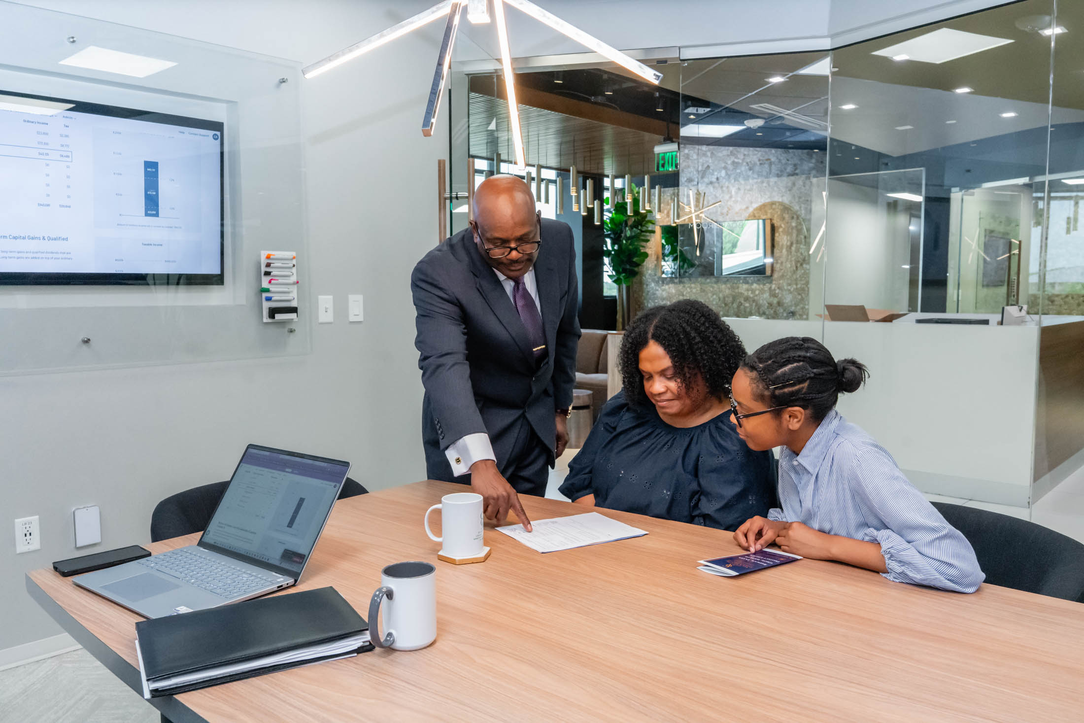 Three people sit at a conference table while a man in a suit points to a document. Laptops, a tablet, and a mug are on the table, with office windows and whiteboard in the background.