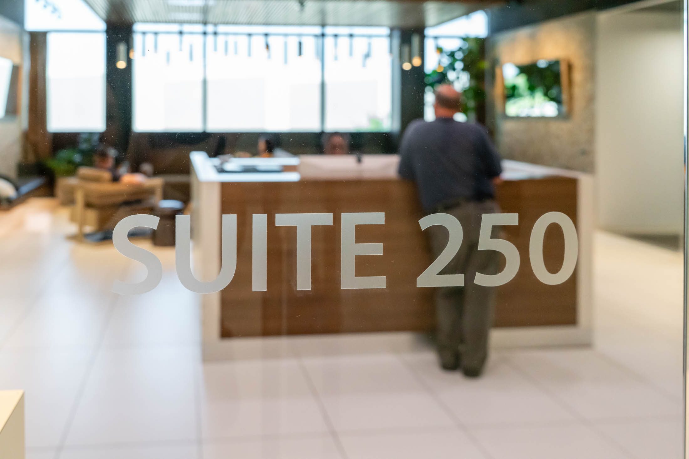 Glass door labeled “Suite 250” with a man standing at a reception desk in a modern office lobby.