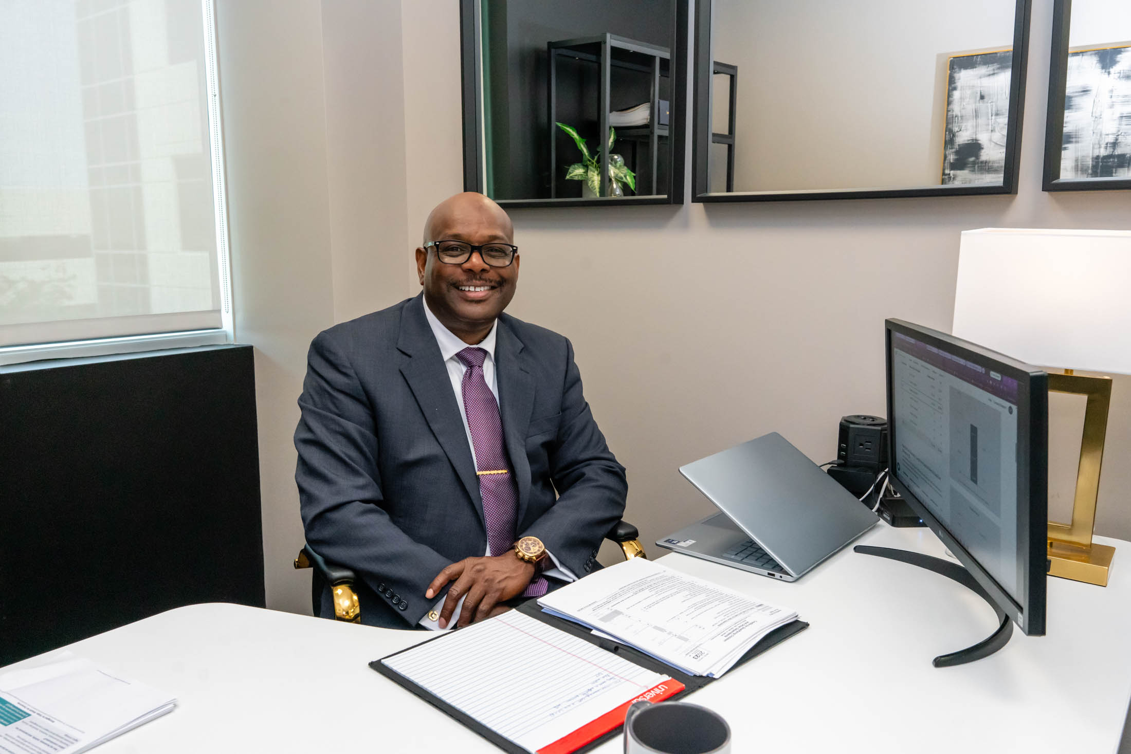 A man in a suit sits at a desk in a modern office, smiling at the camera. A laptop, monitor, documents, and office supplies are on the desk.