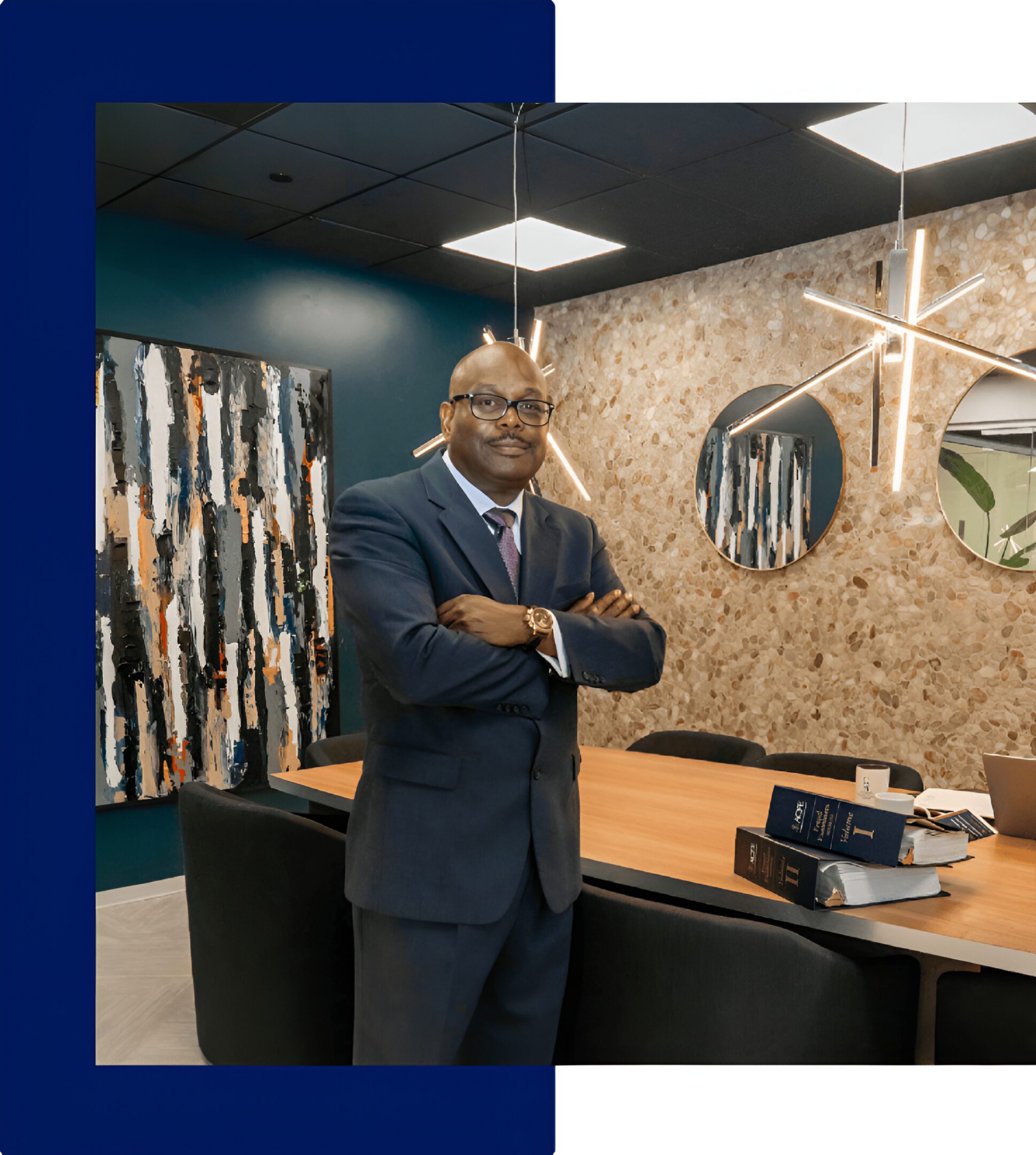Man in a suit stands with arms crossed in a modern office with abstract art, round mirrors, and law books on a wooden table.