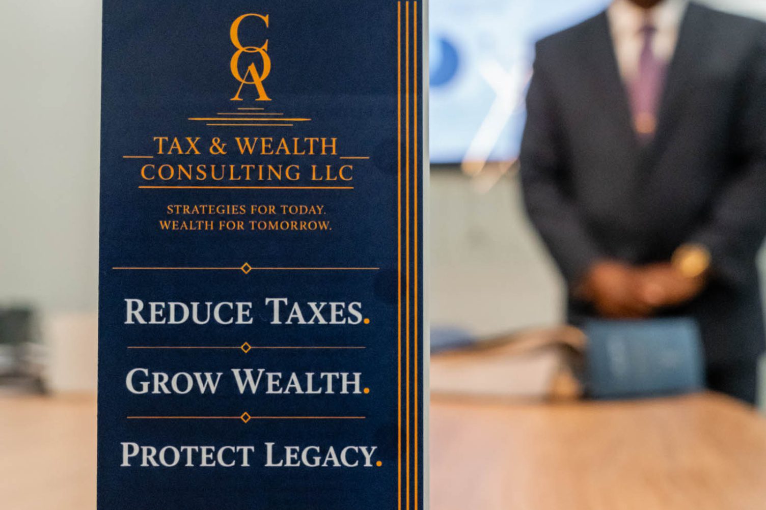 A sign for Tax & Wealth Consulting LLC displays the phrases: Reduce Taxes. Grow Wealth. Protect Legacy. A person in a suit stands blurred in the background.